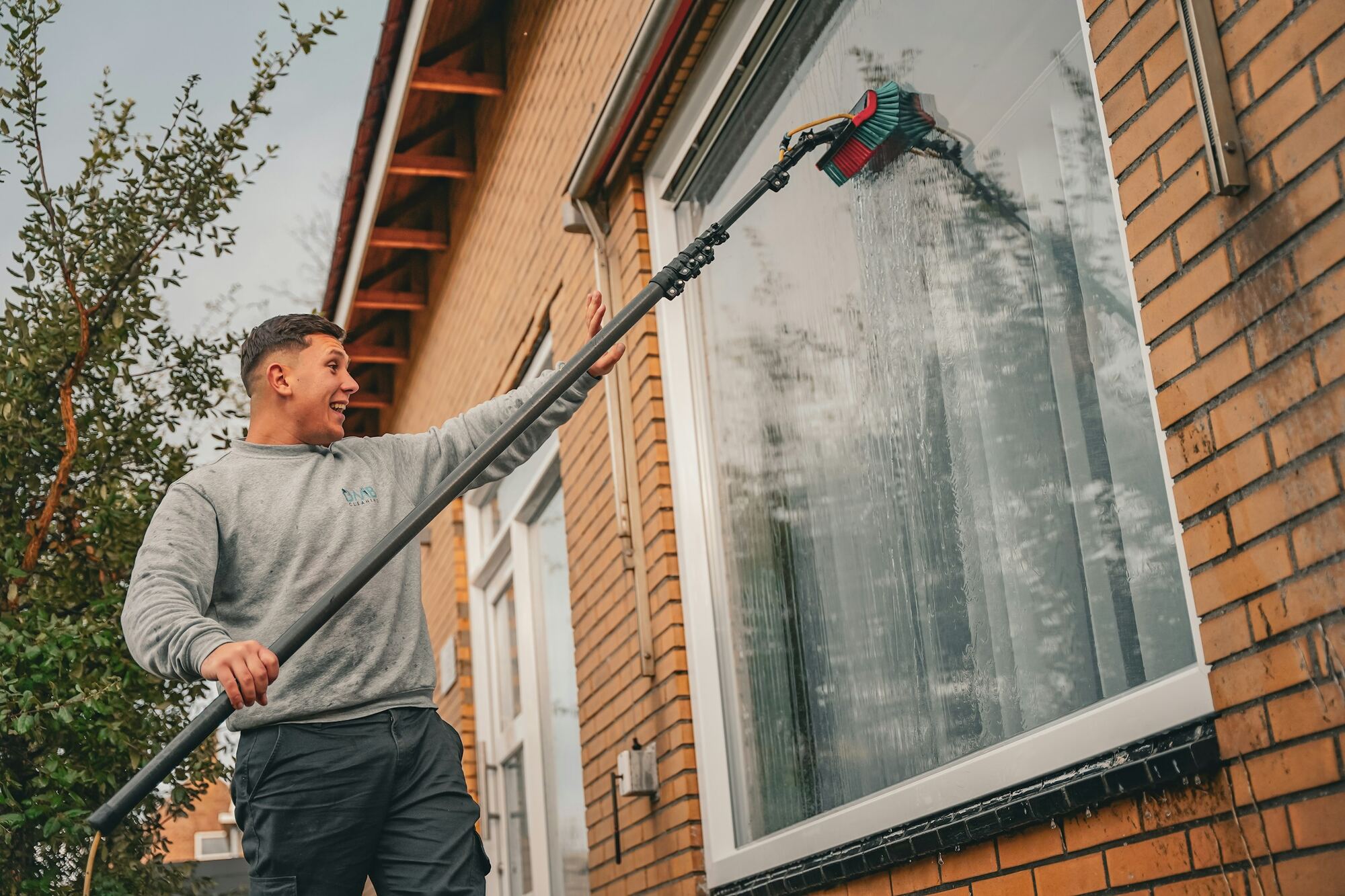 A window cleaner working a water-fed pole across the first-floor windows of a brick house.
