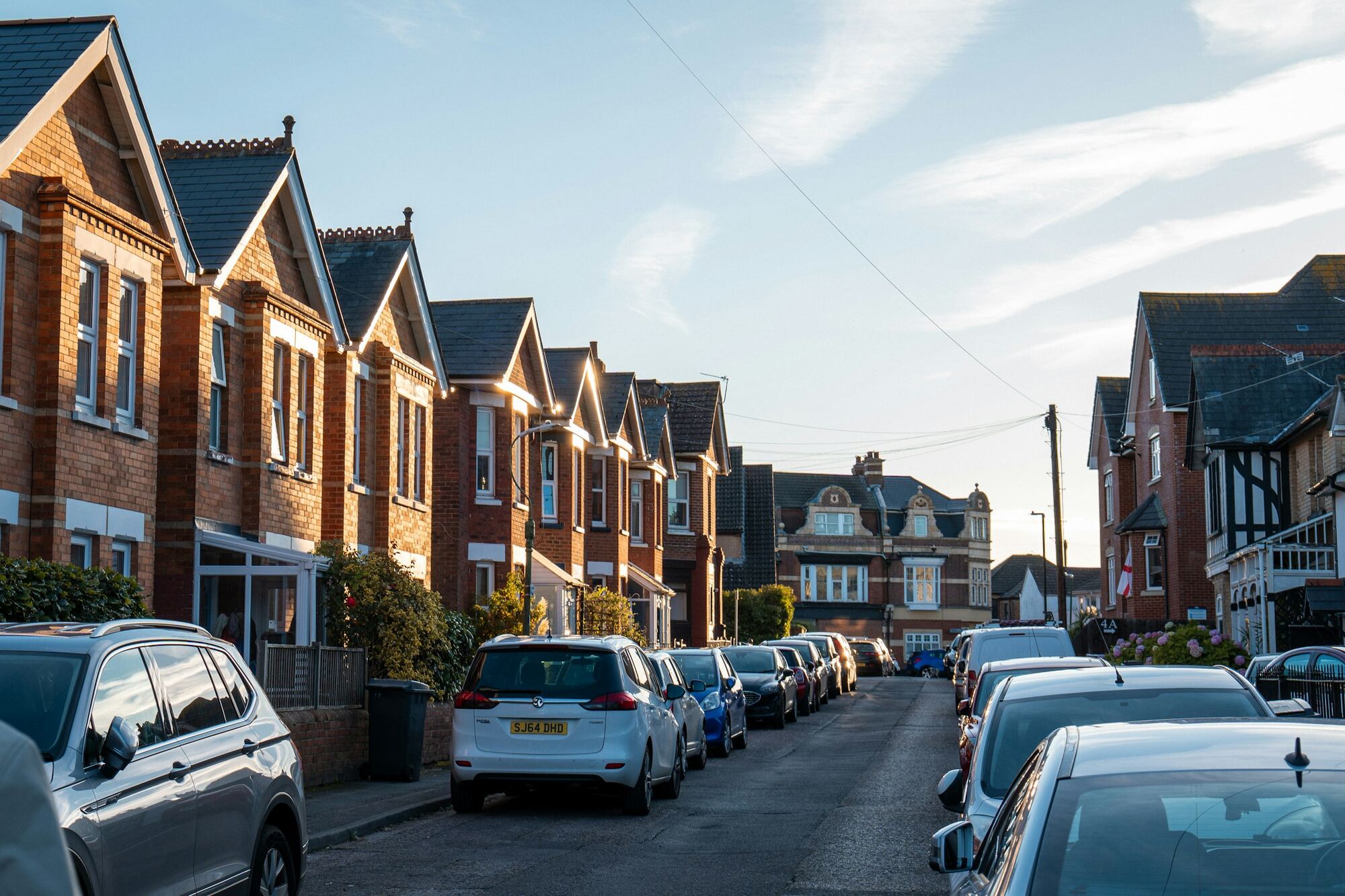 A street of red-brick terraced houses with bay windows in low evening sun, cars parked along both sides.